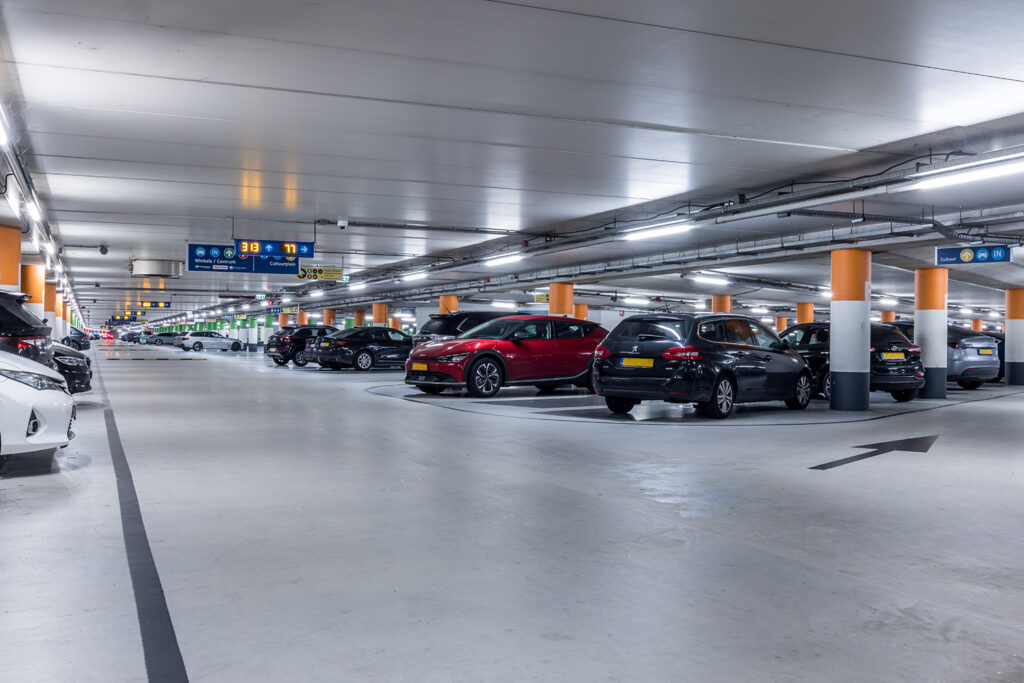 Parkeren Amsterdam Centraal - Hier zie je een afbeelding die is genomen in de parkeergarage van Amsterdam Centrum Parkeren. Je staat in een gangpad in de garage waar je aan de rechterzijde een aantal geparkeerde auto's ziet staan. Je kunt zo de verte in kijken en ziet zo nog heel veel auto's staan